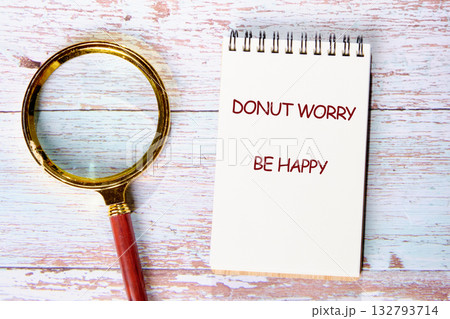 A spiral notebook sits on a wooden surface with the phrase Donut Worry Be Happy written on it. Next to the notebook is a magnifying glass, perhaps searching for joy 132793714