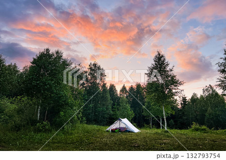 White tourist tent with camping gear in grassy clearing against backdrop of dense trees. Sky glows with vibrant hues of orange and pink as sun sets, casting serene ambiance over peaceful camping site. 132793754