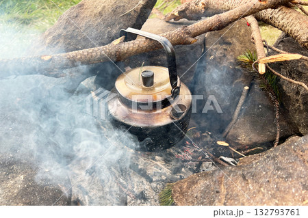 Blackened kettle heating over campfire. Smoke swirls around kettle, and hint of green pine needles visible in foreground. Rustic essence of outdoor cooking. Blackened kettle heating over campfire. Smoke swirls around kettle, and hint of green pine needles visible in foreground. Rustic essence of outdoor cooking. 132793761