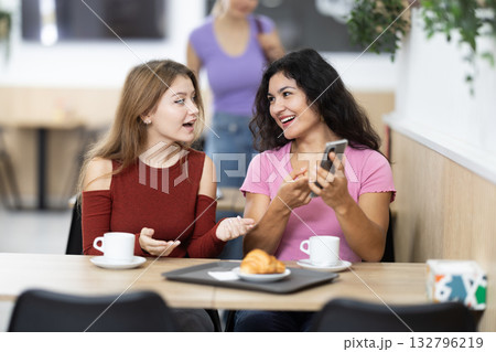 Two positive young girls chatting over coffee and croissant while scrolling smartphone 132796219