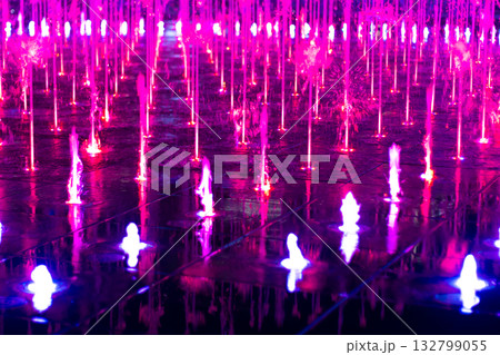 Dancing fountain illuminated pink purple night. Vibrant water jets reflect off a wet, tiled floor, creating an intense, mesmerizing 132799055
