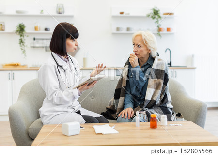A female doctor in a white coat consults with an elderly patient wrapped in a blanket on a sofa, discussing health issues. A female doctor in a white coat consults with an elderly patient wrapped in a blanket on a sofa, discussing health issues. 132805566