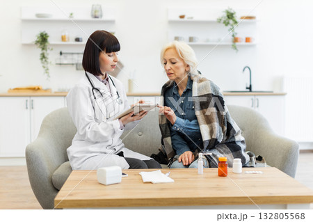 A female doctor in a white coat consults with an elderly patient wrapped in a blanket, discussing health concerns. 132805568