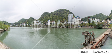 A panoramic view of a Vietnamese town nestled beside a tranquil lake, surrounded by lush green mountains under an overcast sky. 132808730
