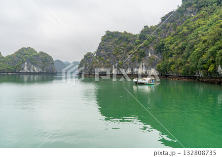 A serene boat floats on the tranquil waters of Ha Long Bay, Vietnam, surrounded by lush limestone karsts. The emerald water mirrors the overcast sky. A serene boat floats on the tranquil waters of Ha Long Bay, Vietnam, surrounded by lush limestone karsts. The emerald water mirrors the overcast sky. 132808735