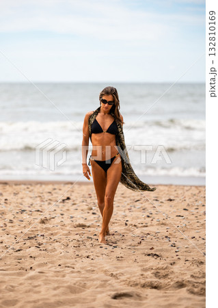 A confident woman strolls along the beach, showcasing her vibrant summer style and relaxation vibes 132810169