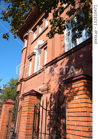 Facade of an old red brick building with arched windows and decorative white accents, partially shaded by trees and sunlight reflections. The image is suitable for topics about building restoration Facade of an old red brick building with arched windows and decorative white accents, partially shaded by trees and sunlight reflections. The image is suitable for topics about building restoration 132810471