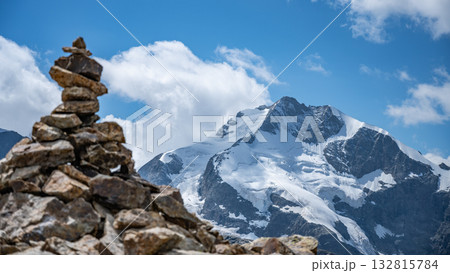 A stone cairn stands prominently in the foreground against the majestic backdrop of Piz Bernina in the Swiss Alps. The glacier-covered mountain towers under a bright blue sky. A stone cairn stands prominently in the foreground against the majestic backdrop of Piz Bernina in the Swiss Alps. The glacier-covered mountain towers under a bright blue sky. 132815784