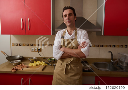 Male Cook in Beige Apron Stands Confidently in Red Kitchen While Preparing Potatoes Male Cook in Beige Apron Stands Confidently in Red Kitchen While Preparing Potatoes 132820190