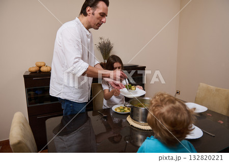 Father Cooks Soup For His Kids While They Eat Dinner At Home And Help With Plate Father Cooks Soup For His Kids While They Eat Dinner At Home And Help With Plate 132820221