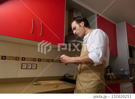 Man In Beige Apron Preparing Dinner In A Modern Kitchen With Red Cabinets Man In Beige Apron Preparing Dinner In A Modern Kitchen With Red Cabinets 132820251