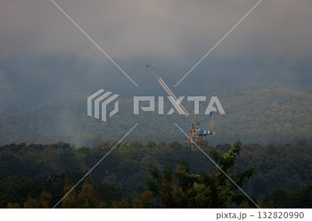Industrial Marvel Above The Clouds: A towering crane pierces the misty atmosphere above a dense forest, symbolizing progress, industry, and the fusion of human endeavor with nature. 132820990