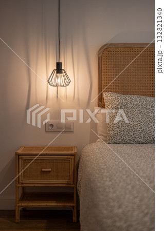 A cozy bedroom scene featuring a bed with a patterned white duvet and a rattan headboard. A bedside table holds a stylish pendant lamp, creating a warm ambiance. 132821340