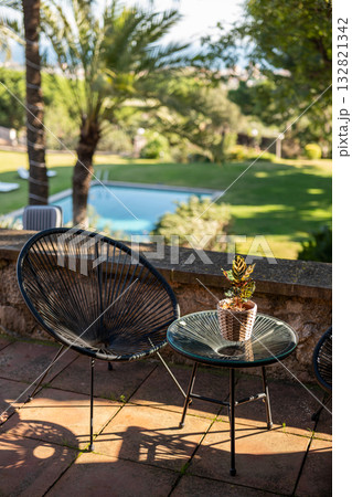 A black chair and a glass table with a potted plant are set on a patio overlooking a green lawn and swimming pool. Palm trees are visible in the background. 132821342