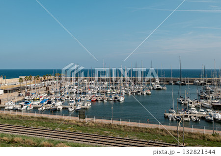 A marina in Barcelona with numerous sailboats docked. The clear blue sky reflects on the calm water. Green grass and railway tracks are visible nearby. A marina in Barcelona with numerous sailboats docked. The clear blue sky reflects on the calm water. Green grass and railway tracks are visible nearby. 132821344