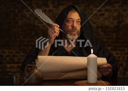 A hooded man with a quill in his hand is immersed in work on a manuscript. 132821813