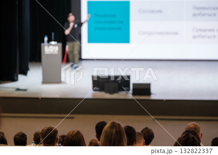 Business conference presentation on stage with speaker presenting to audience and large screens Business conference presentation on stage with speaker presenting to audience and large screens 132822337