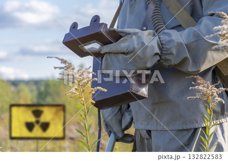A military man in chemical protection and a gas mask measures the radiation level with a dosimeter in nature. Concept: radiation leakage, radioactive hazard. 132822583