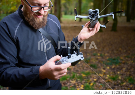 A man enjoys flying a drone in a vibrant green forest. He carefully checks the remote control as sunlight filters through the trees, creating a serene atmosphere. 132822816