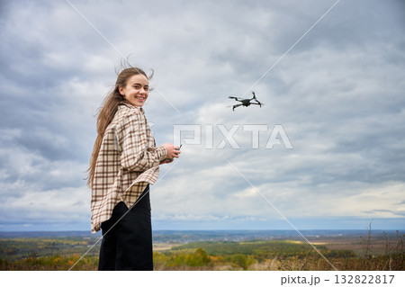 A girl stands on a mountain top, smiling as she controls a drone. Wisps of her hair dance in the wind, and the sky is filled with clouds, creating a serene atmosphere. 132822817