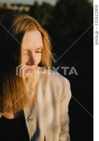Young blonde woman with closed eyes enjoying warm light of sunset in natural environment, wearing light linen jacket and black shirt. Portrait of woman in linen jacket and black shirt, eyes closed 132822900