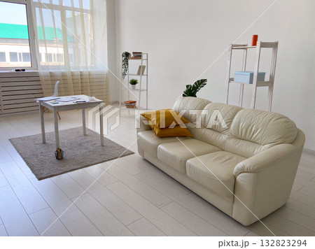 Bright modern interior with cream sofa, yellow pillows, desk, and shelving units near large window. Calm atmosphere, simplicity, and Scandinavian inspired home decor. Bright modern interior with cream sofa, yellow pillows, desk, and shelving units near large window. Calm atmosphere, simplicity, and Scandinavian inspired home decor. 132823294