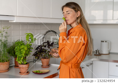 Woman gardener enjoys growing aromatic herbs at home, holding scissors and smelling leaf of basil. 132824954