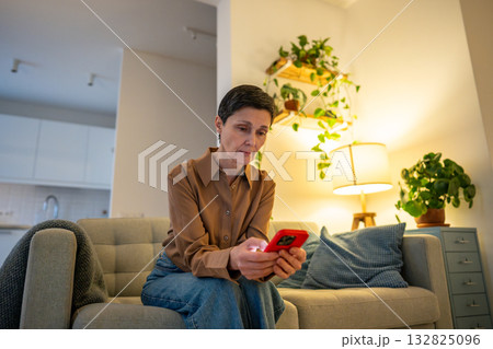 Mature woman scrolls endlessly in smartphone lost in digital distraction at home, scrolling numbing 132825096