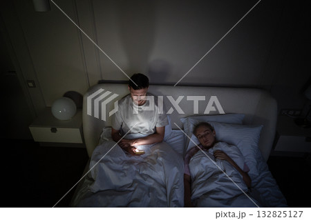 Exhausted man awake alone in bed scrolling phone due to insomnia and digital addiction on late hours 132825127