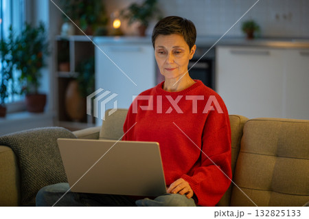 Tranquil mature woman work calmly on laptop on sofa in comfortable living room. Female productivity 132825133