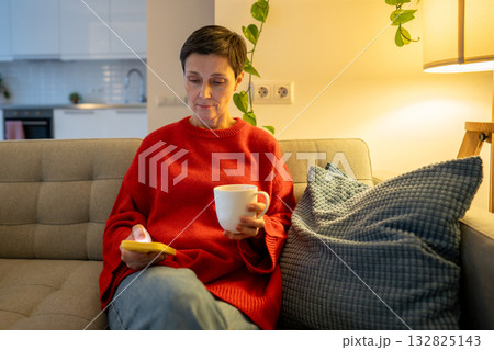 Thoughtful mature woman scrolling smartphone. Reflective female drinking tea on evening sofa Thoughtful mature woman scrolling smartphone. Reflective female drinking tea on evening sofa 132825143