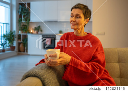 Calm mature woman drinking hot tea on sofa lost in thought in home comfort. Old female reflection Calm mature woman drinking hot tea on sofa lost in thought in home comfort. Old female reflection 132825146
