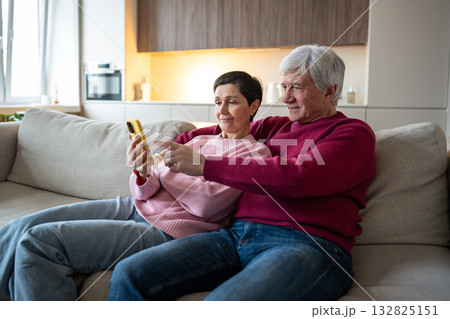 Pleased elderly couple on sofa scrolling social media reels on shared smartphone, social media enjoy 132825151