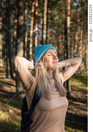 Middle-aged woman practicing mindfulness, turning face to sun during walk in pine forest wilderness  132825224