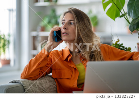 Stunned middle-aged woman receiving sudden shocking news via smartphone while seated at laptop Stunned middle-aged woman receiving sudden shocking news via smartphone while seated at laptop 132825227