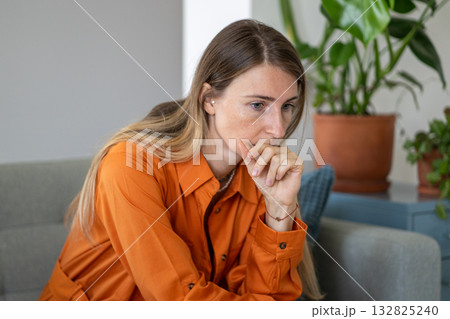 Worried middle-aged woman touching her chin deeply absorbed in thought sitting alone on sofa Worried middle-aged woman touching her chin deeply absorbed in thought sitting alone on sofa 132825240