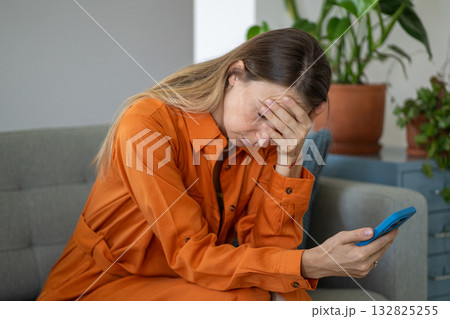 Shocked middle-aged woman in distress holding head with hand and smartphone seated on home sofa Shocked middle-aged woman in distress holding head with hand and smartphone seated on home sofa 132825255