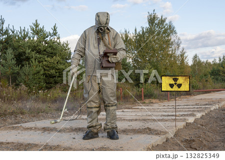 A military man in chemical protection and a gas mask measures the radiation level with a dosimeter, a radiation hazard sign, a concept: radiation leakage, the threat of infection. A military man in chemical protection and a gas mask measures the radiation level with a dosimeter, a radiation hazard sign, a concept: radiation leakage, the threat of infection. 132825349