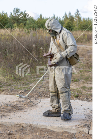 A military man in chemical protection and a gas mask measures the radiation level with a dosimeter in nature, smoke from a fire. Concept: radiation leakage, radioactive hazard. A military man in chemical protection and a gas mask measures the radiation level with a dosimeter in nature, smoke from a fire. Concept: radiation leakage, radioactive hazard. 132825427