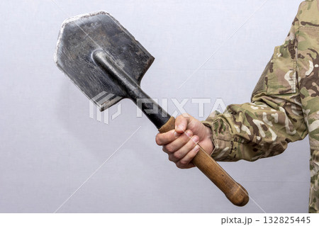 A soldier in an American military uniform holds a sapper shovel in his hands on a light background. A soldier in an American military uniform holds a sapper shovel in his hands on a light background. 132825445
