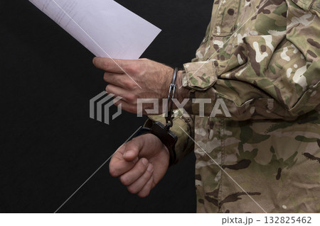 a military man in a camouflage uniform is handcuffed on a black background, with a white sheet of paper in his hand. a military man in a camouflage uniform is handcuffed on a black background, with a white sheet of paper in his hand. 132825462