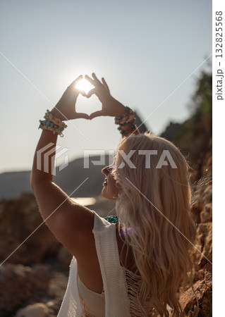 Woman, sun, heart. Blonde woman making a heart shape with hands against setting sun on a rocky beach, radiating peace and connection Woman, sun, heart. Blonde woman making a heart shape with hands against setting sun on a rocky beach, radiating peace and connection 132825568