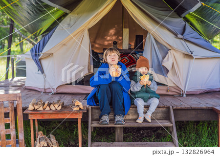Toddler and his teenage brother tourists in warm vests and hats eating buns while sitting on glamping steps, enjoying family travel, outdoor adventure, and bonding Toddler and his teenage brother tourists in warm vests and hats eating buns while sitting on glamping steps, enjoying family travel, outdoor adventure, and bonding 132826964