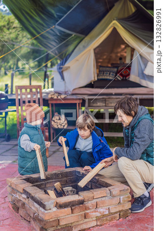 Father and his two sons making a campfire in front of a glamping site, enjoying family bonding, outdoor adventure, and nature exploration 132826991