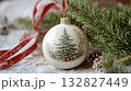 A delicate round ceramic ornament with a hand-painted Christmas tree, surrounded by intricate pine needles and subtle holiday accents. 132827449