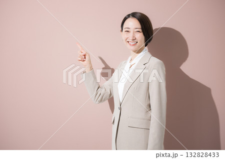 ピンク背景でカメラ目線で立つスーツの女性　オススメや紹介のポイントの指差しポーズ 132828433