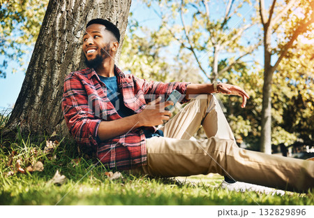 Nature, phone and man sitting by a tree in an outdoor park networking on social media or the internet. Technology, rest and African guy browsing on mobile app with cellphone while relaxing in garden. 132829896