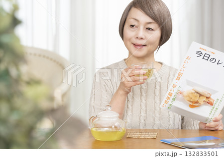 夏リビングで冷茶を飲みながらレシピ雑誌を見るシニア女性 132833501