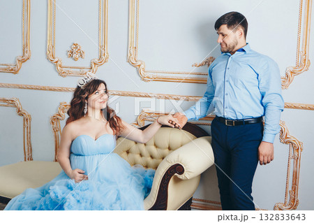 a married couple stands together near a beige sofa. couple in love looking at each other. pregnant girl in a lush blue dress. young man in a blue shirt. family photo session. a married couple stands together near a beige sofa. couple in love looking at each other. pregnant girl in a lush blue dress. young man in a blue shirt. family photo session. 132833643