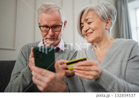 Senior couple man woman shopping online with smartphone paying with credit card. Old people buying on Internet enter credit card details at home indoors. Online shopping delivery service ecommerce Senior couple man woman shopping online with smartphone paying with credit card. Old people buying on Internet enter credit card details at home indoors. Online shopping delivery service ecommerce 132834537